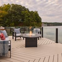 A serene outdoor deck featuring a modern fire pit surrounded by cushioned chairs. The setting overlooks a calm lake with lush green trees in the background under a cloudy sky. Cozy blankets and pillows add a touch of comfort to the seating arrangement.